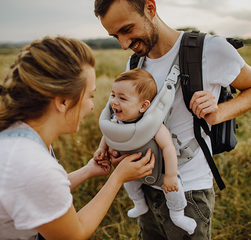A couple adjusts a baby carrier on a man's chest, with the baby comfortably seated inside, set against a scenic outdoor backdrop of fields and distant trees.
