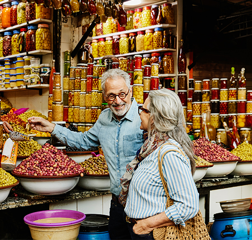 A retired couple, including a woman with long grey hair and wearing a striped shirt and a man wearing a denim shirt, browse a vibrant market stall filled with colorful jars and bowls of preserved fruits and vegetables.