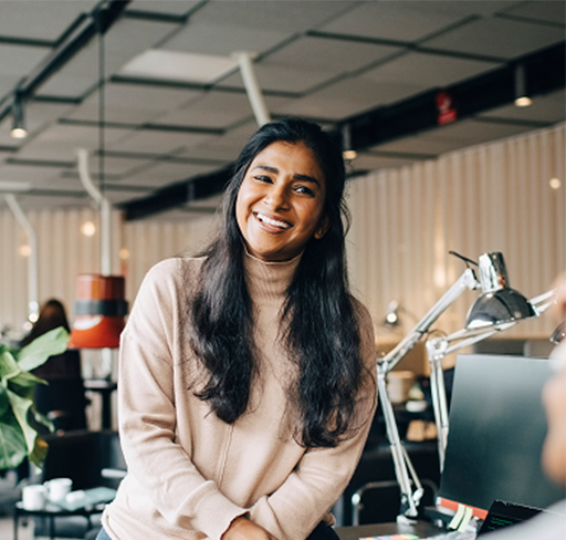 A person with long dark hair, wearing a light beige sweater, is leaning slightly forward at a desk in a modern, well-lit office space with contemporary decor and desk lamps.