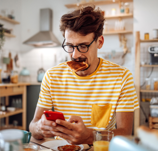 A person wearing a yellow and white striped shirt sits at a table in a kitchen, looking down at a mobile phone while holding a piece of food in their mouth, with cups, a glass of juice, and kitchen shelves in the background.