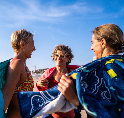 Three adult women standing together at a beach, holding patterned towels with water and sky behind them.