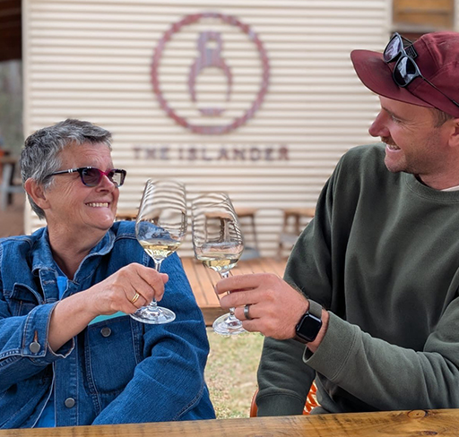Two people sit at a wooden outdoor table, clinking glasses of white wine together at a winery, with a rustic building in the background.