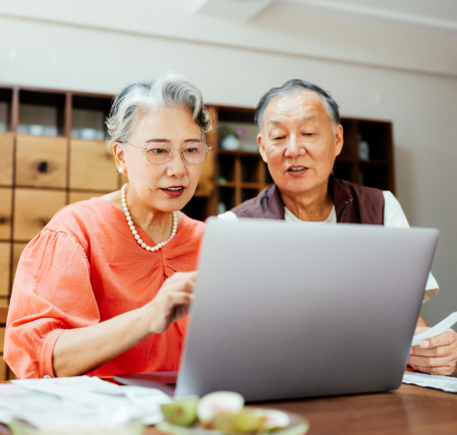 Two older adults review documents together while using a laptop at a table in a home office setting.