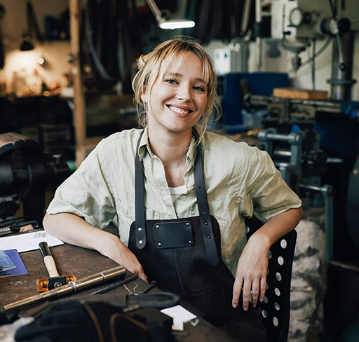 A woman wearing a light-colored shirt and dark apron sits at a workbench in a workshop, surrounded by various tools and machinery.