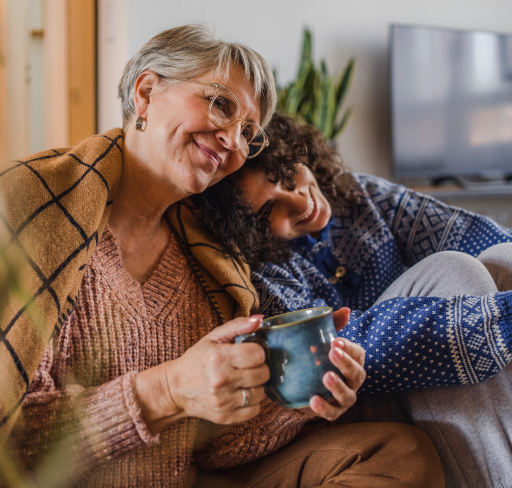 An older woman and a younger woman sit together on a couch at home, sharing a quiet moment with a warm drink.