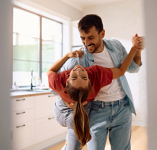 A man playfully dips a woman backward in a dance pose in a bright, modern kitchen. She wears a red t-shirt, and he is in a light blue shirt over a white t-shirt with blue jeans.