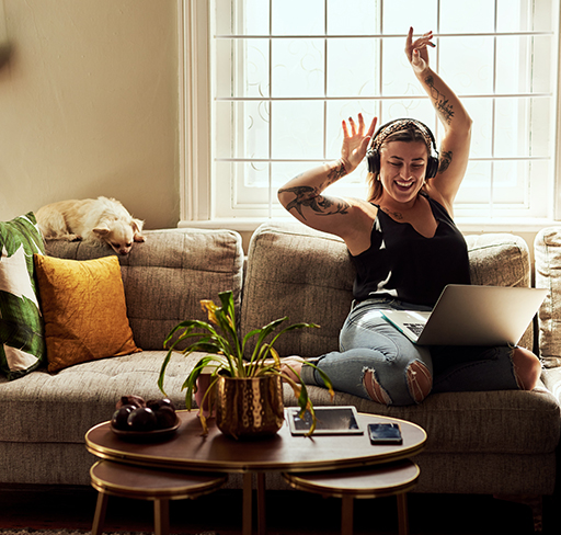 Person sitting on a couch with a laptop and headphones, raising one arm while relaxing in a sunlit living room. A small dog rests on the couch nearby and a coffee table with plants and a tablet is in the foreground.
