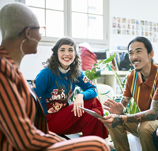 A group of four people are having a conversation in a bright office space, with one person gesturing while holding a tablet, surrounded by modern decor and large windows.