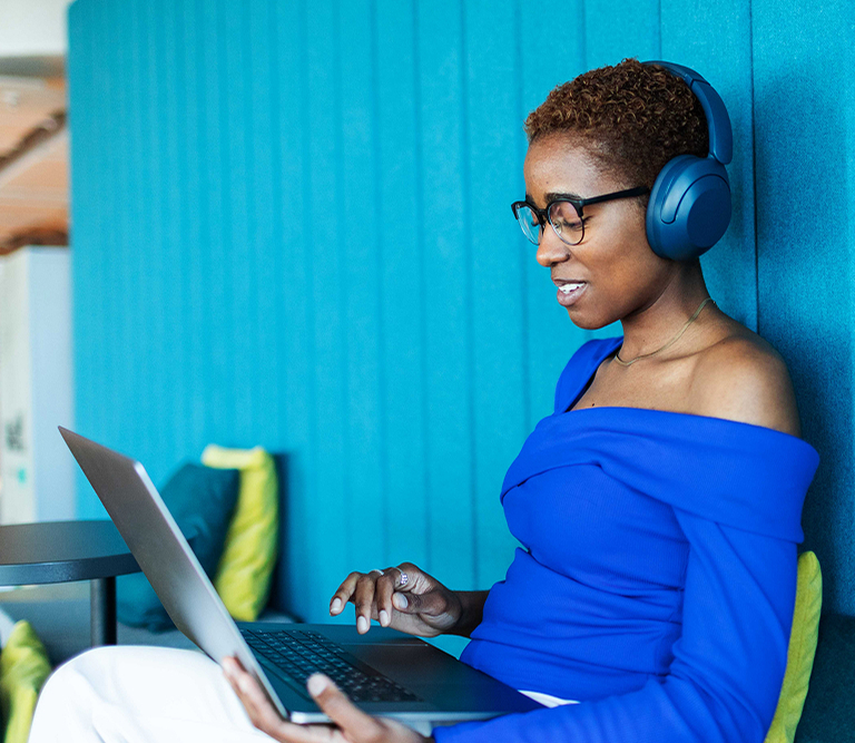 Woman wearing headphones, leaning against blue wall, holding and typing on laptop