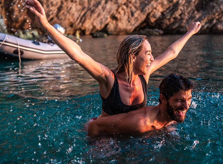 A couple riding piggyback in shallow water near a rocky shore