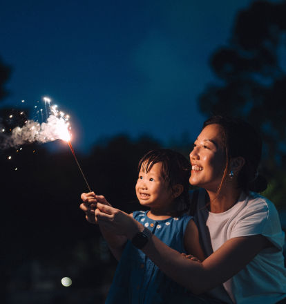 A woman in a white shirt holds a sparkler with her smiling child.