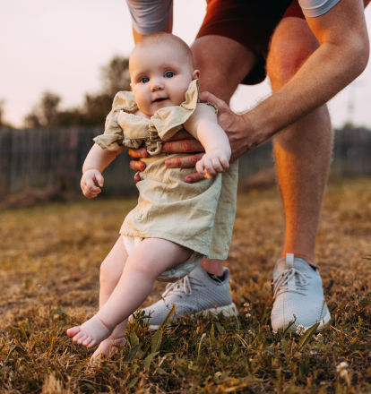 A person in white sneakers holds a smiling baby above a grass lawn.